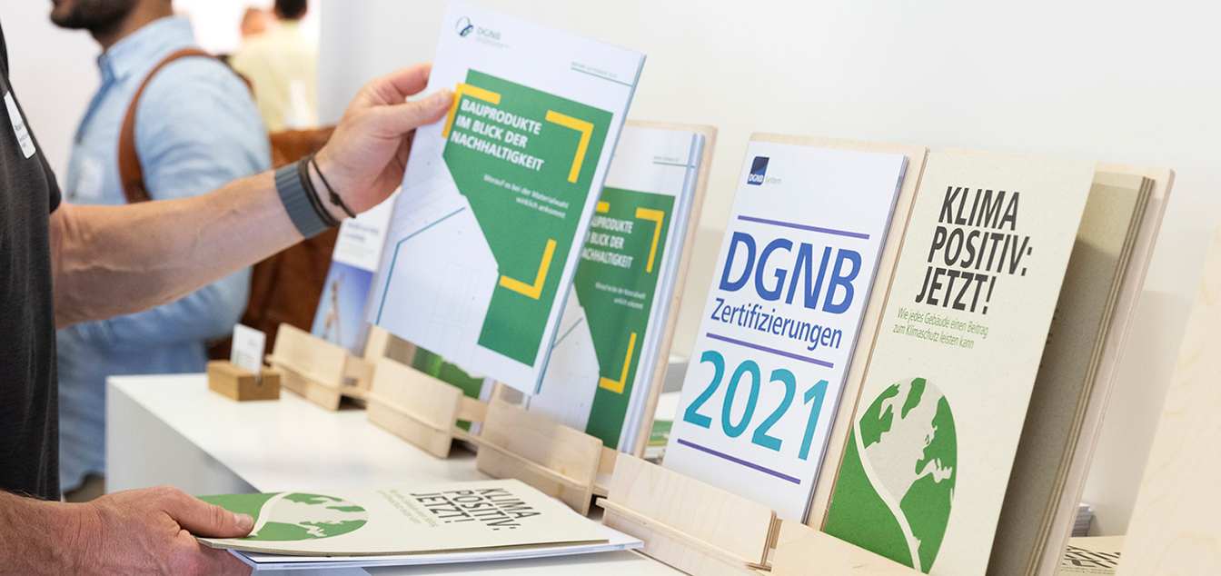 Eine Hand nimmt vom linken Bildrand aus eine Broschüre zum Thema Bauprodukte aus einem Aufsteller. Weitere Broschüren sind ausgestellt.