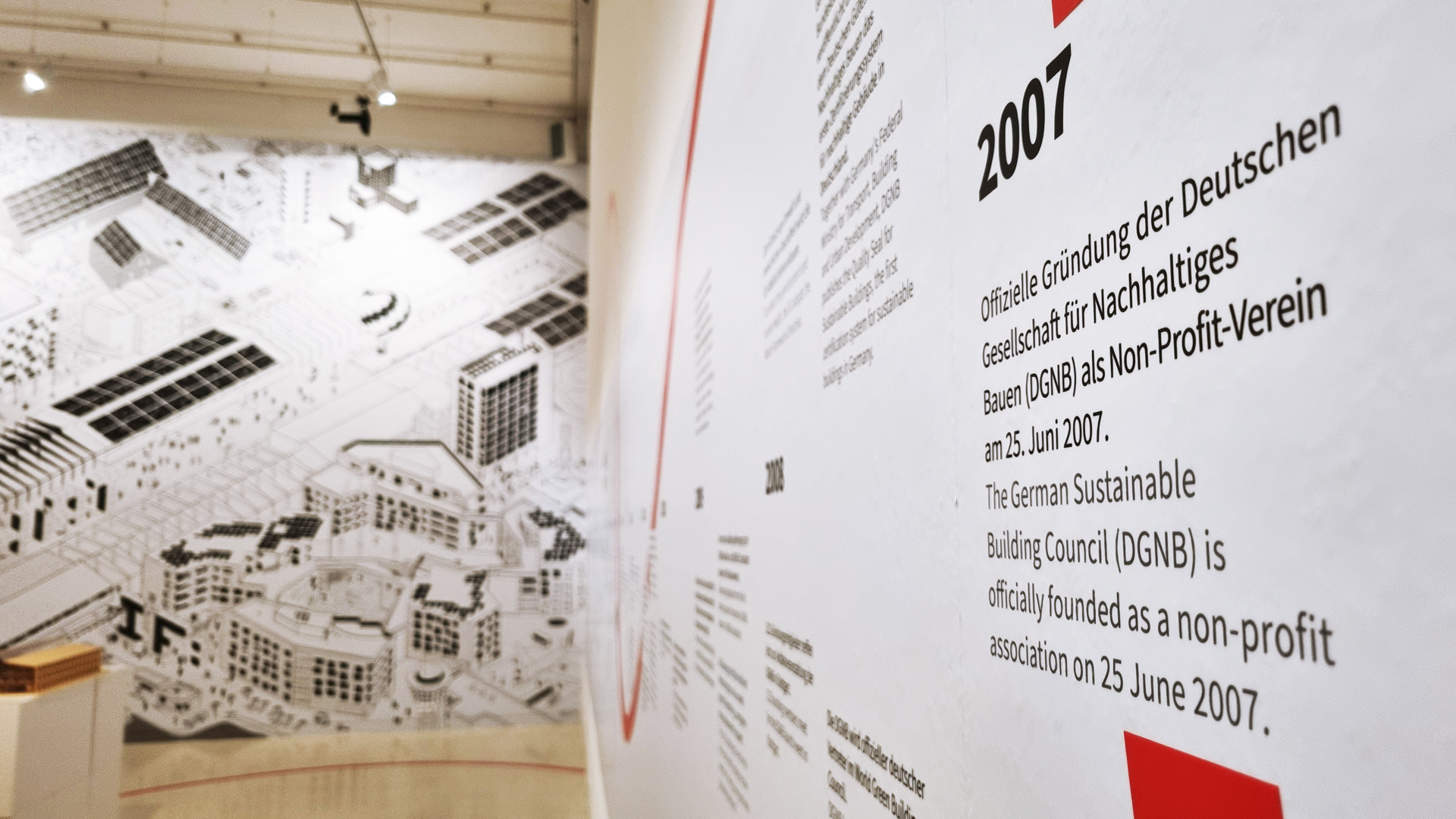 DGNB Ausstellung bei Aedes Das Bild zeigt einen Ausschnitt der DGNB Ausstellung bei Aedes. Zu sehen ist auf der rechten seite ein angeschnittener Zeitstrahl. Der Blick in den Raum gibt die Sicht auf ein großes schwarz-weißes Wimmelbild frei.