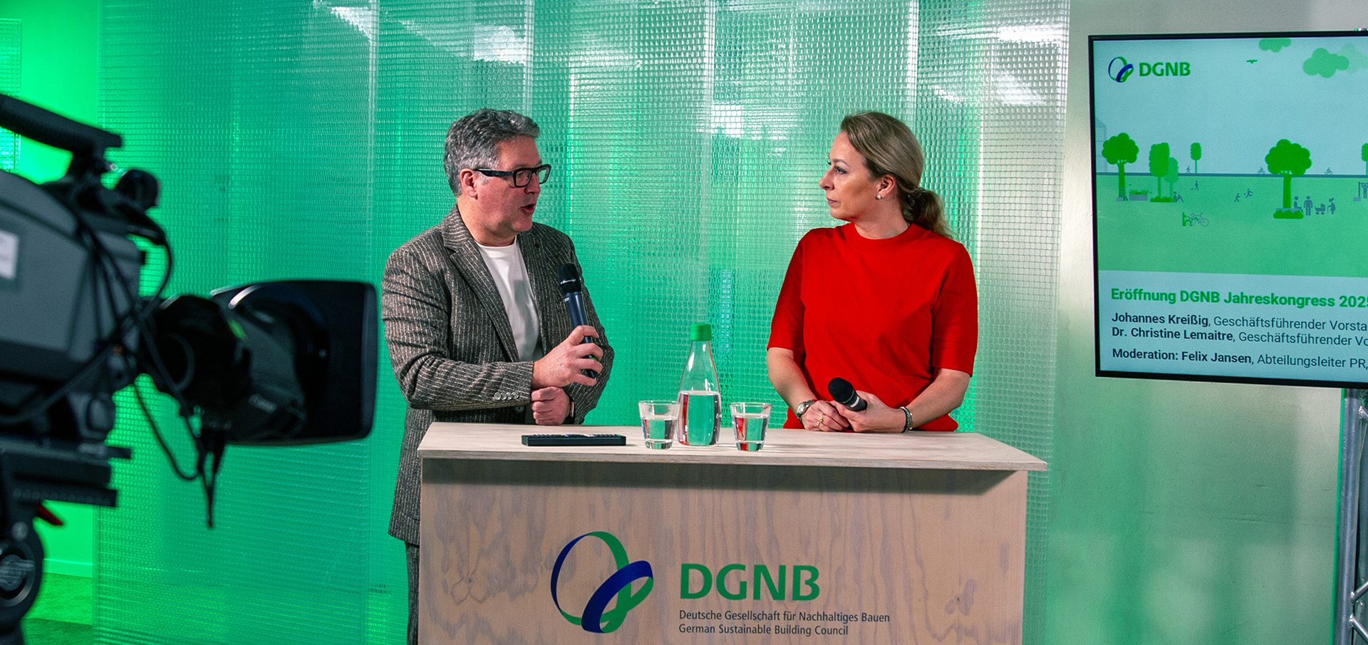 Gesprächssituation mit einer Frau rechts und einem Mann links vor einem podium. Eine Kamera ist am linken Bildrand zu erkennen.