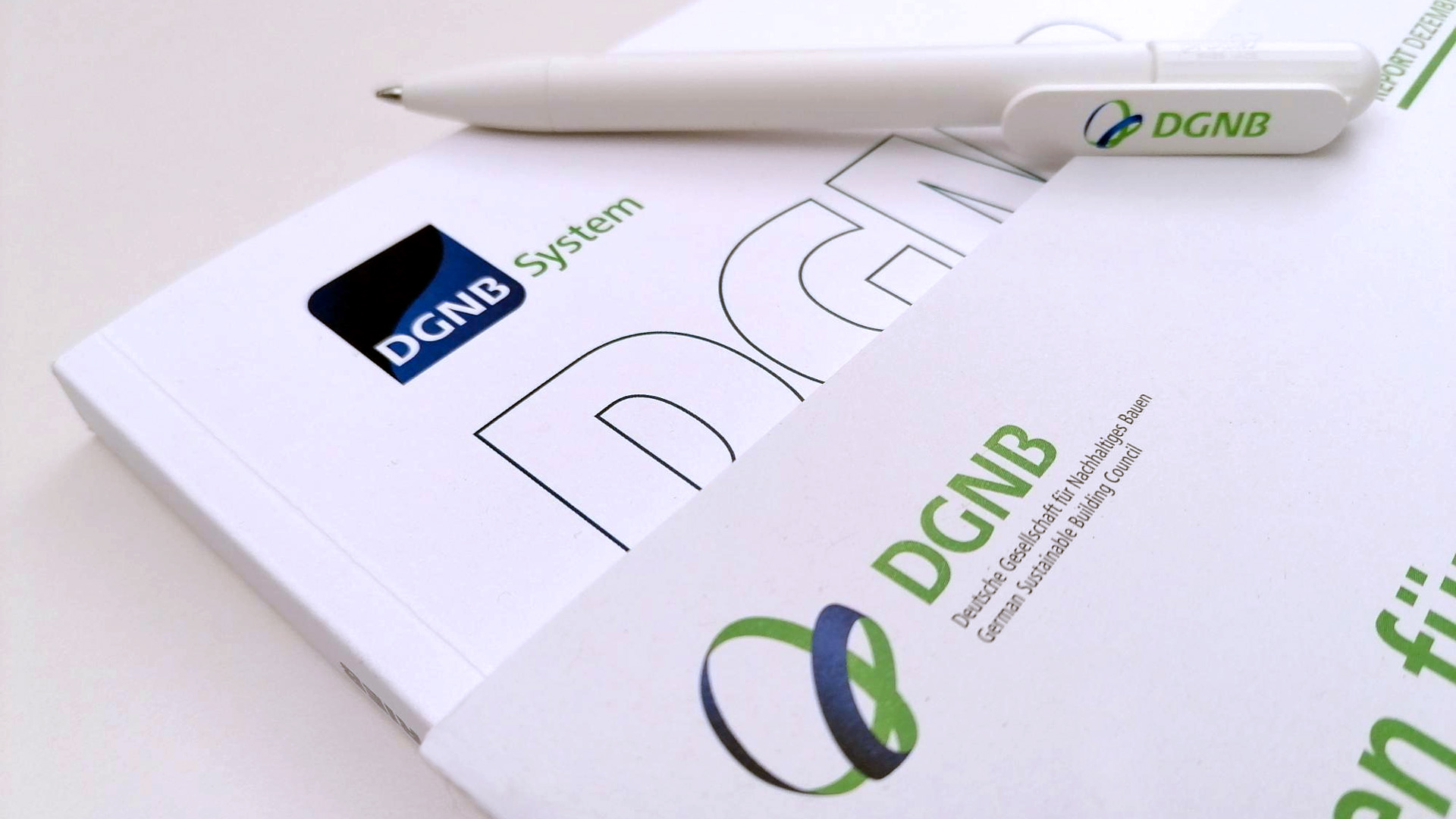Auf dem Foto ist auf weißem Hintergrund ein Ausschnitt von einem DGNB Kriterienkataloog gesehen. Man sieht die obere Ecke mit dem System Logo sowie einem DGNB Schriftzug. Auf dem Katalog liegt ein weiteres Dokument auf dem das Vereinslogo der DGNB abgebildet ist. Auf den Publikationen liegt zude ein weißer Kugelschreiber ebenfalls mit DGNB Logo.