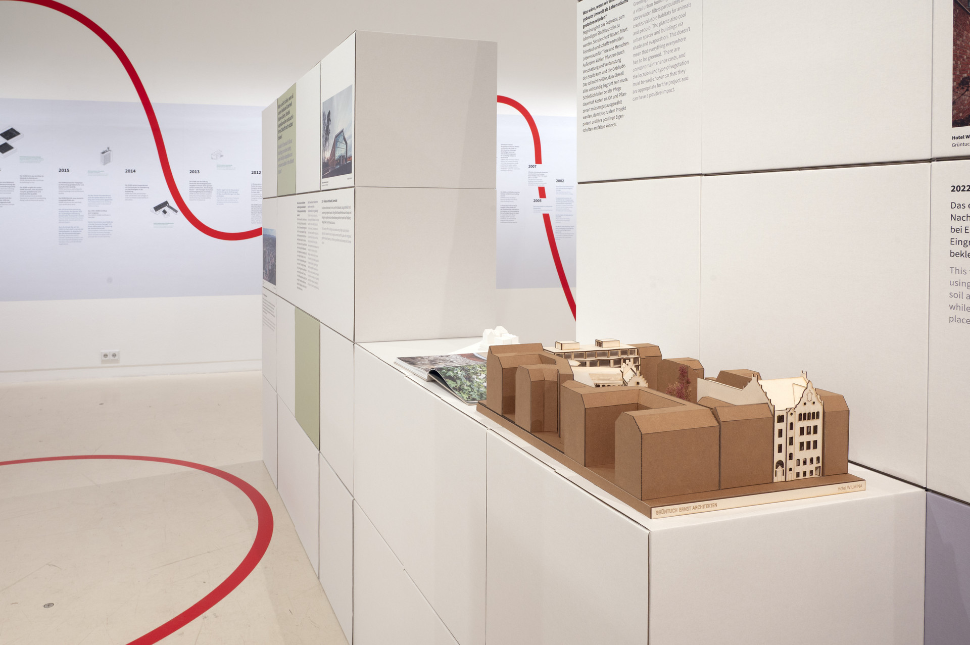 Einblick in die DGNB Ausstellung bei Aedes. Zu sehen sind Ausstellungswände aus Pappwürfeln, eine braunes Gebäudemodell auf einem Sockel. Eine geschwungene rote Linie zieht sich durch den weißen raum. Im Hintergrund ist an der Wand der große Zeitstrahl zu erkennen.