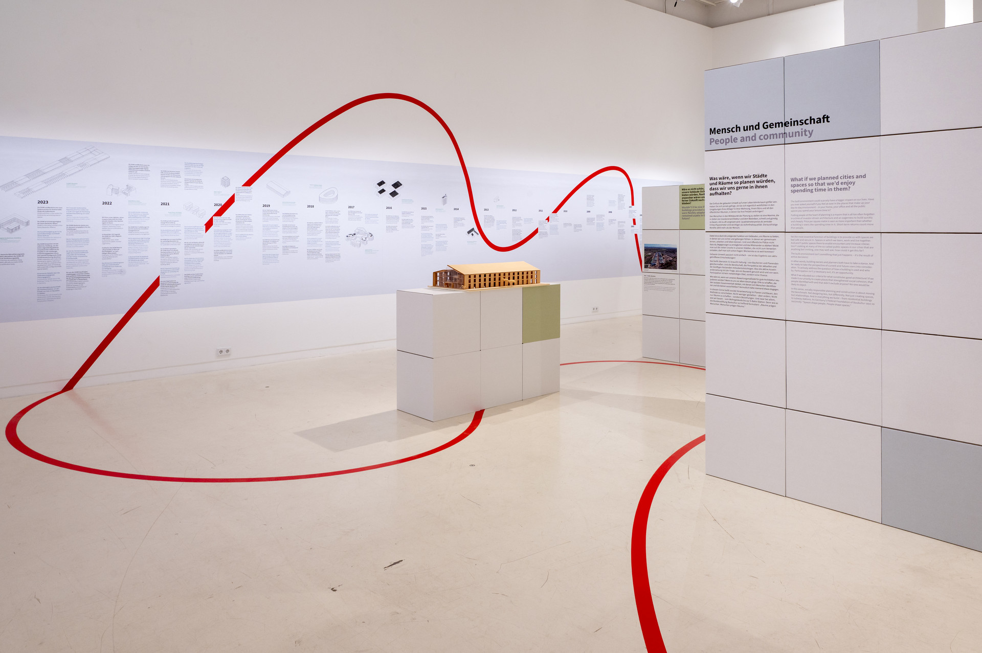 Einblick in die DGNB Ausstellung bei Aedes. Zu sehen sind Ausstellungswände aus Pappwürfeln, eine braunes Gebäudemodell auf einem Sockel. Eine geschwungene rote Linie zieht sich durch den weißen raum. Im Hintergrund ist an der Wand der große Zeitstrahl zu erkennen.
